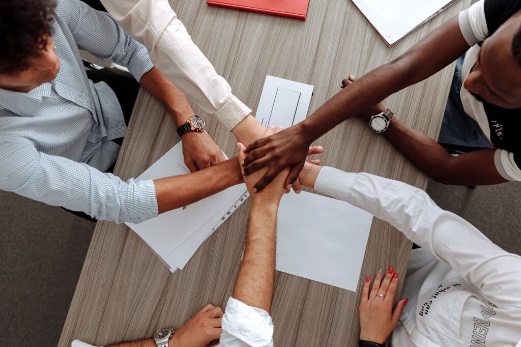 A diverse group of adults join hands in an office, symbolizing teamwork and cooperation.